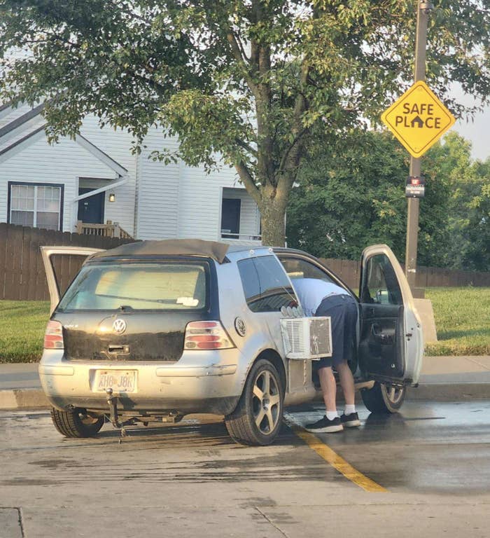 A person is bent over, looking inside the open door of a Volkswagon car in a parking lot. A "Safe Place" sign is visible on a light post nearby