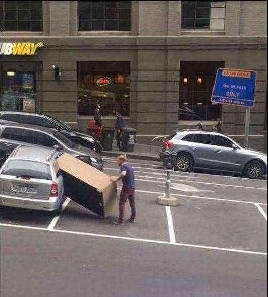 Person attempts to fit a large cardboard box into a car's back seat in a parking lot near several buildings
