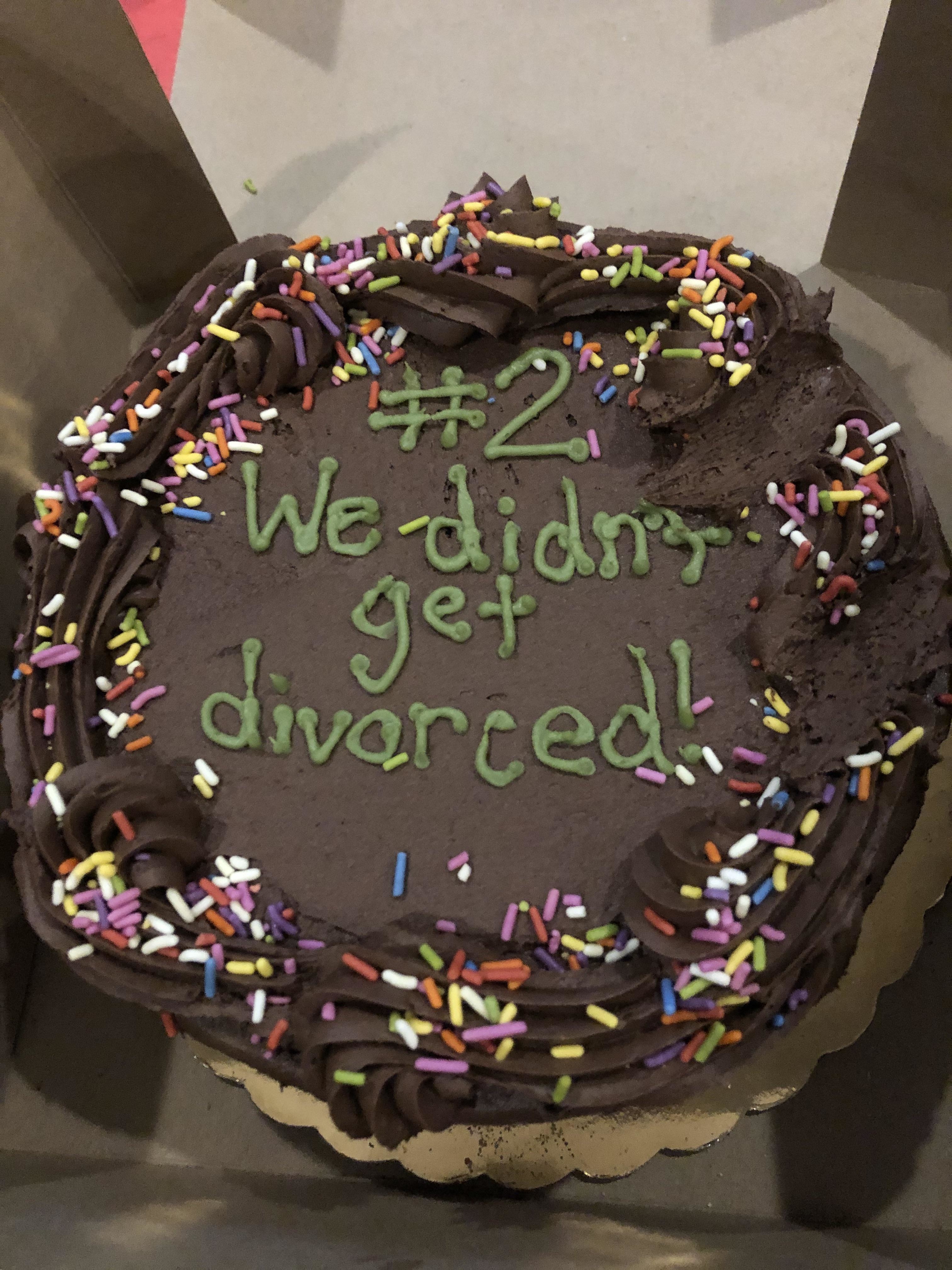 A chocolate cake with colorful sprinkles and damaged frosting. The cake reads "#2. We didn't get divorced!" in green icing