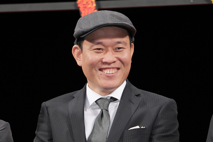 帽子をかぶり、スーツを着た男性がマイクを持ち笑顔で立っています。