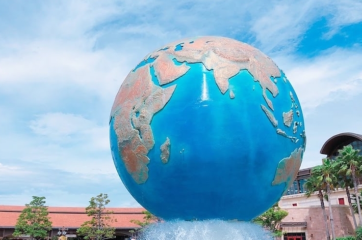 大きな地球儀が噴水の上に設置されているテーマパークのエントランス。背景には青空と植栽、建物が見える。