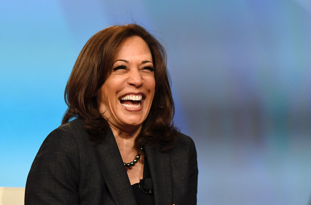 Kamala Harris smiling and laughing while wearing a dark jacket and a pearl necklace in a brightly lit setting