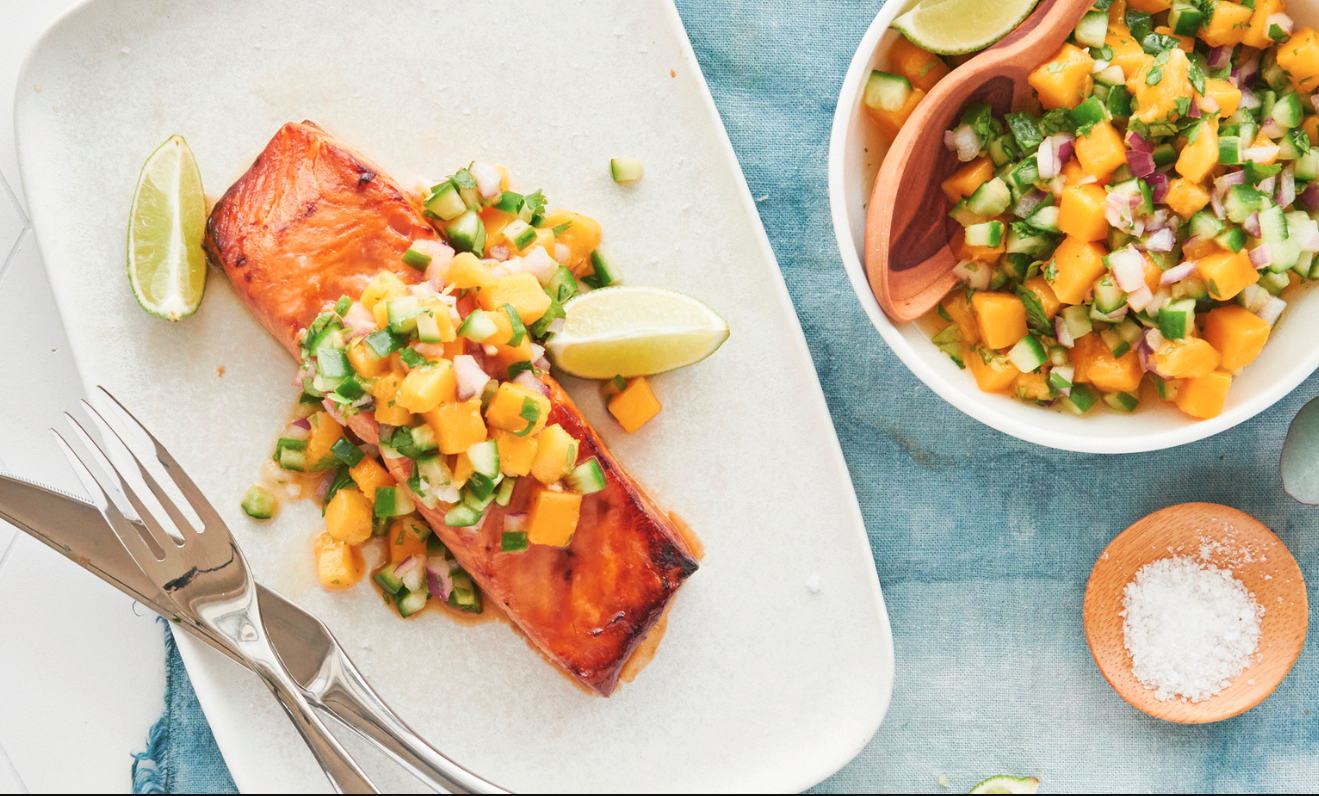 Grilled salmon fillet garnished with a mango salsa, lime wedges, and a bowl of the same salsa on the side, with salt and utensils nearby