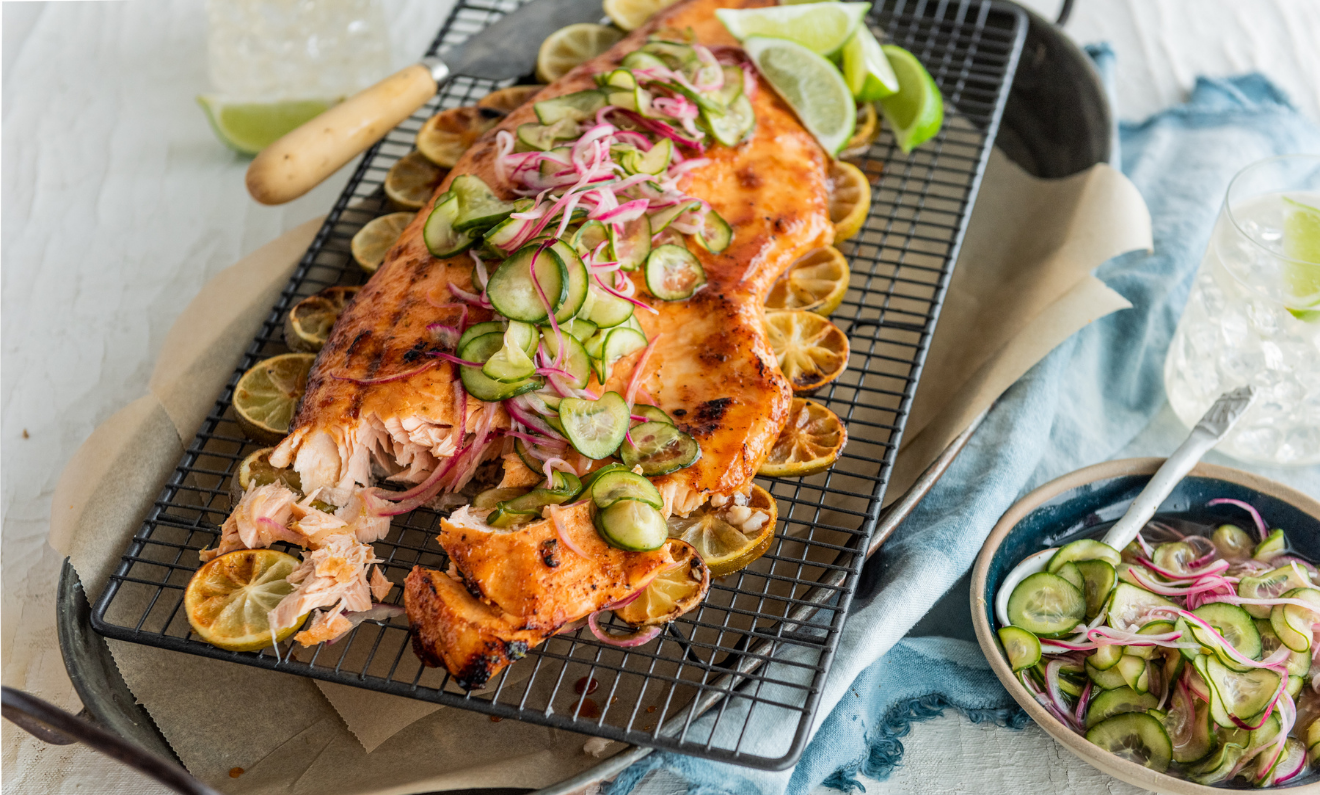 Grilled salmon topped with sliced cucumbers and red onions is served on a bed of lime slices, presented on a rectangular wire rack with lime wedges and a side salad
