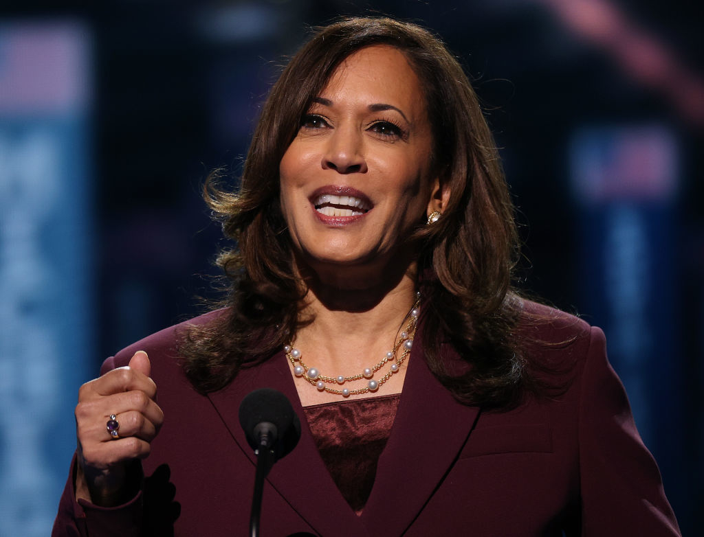 Kamala Harris is speaking into a microphone, wearing a blazer and a pearl necklace