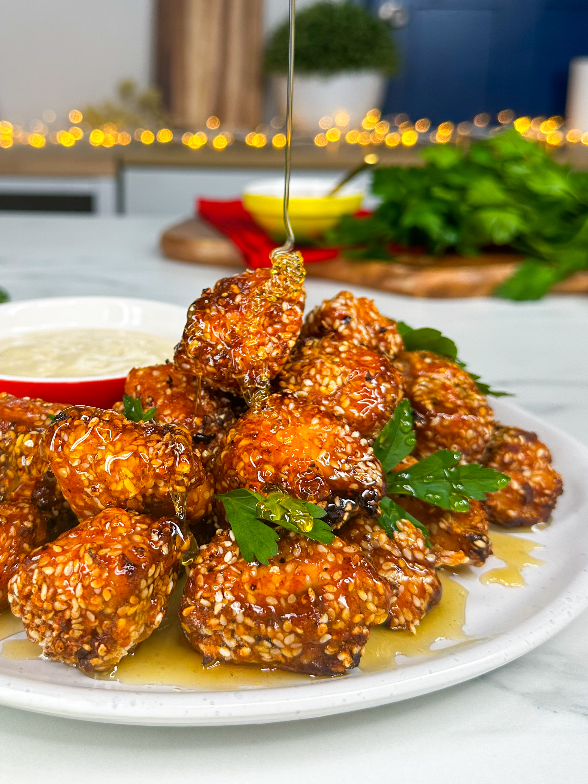 A plate of crispy sesame-covered chicken bites is being drizzled with honey. The background features a bunch of fresh parsley and kitchen items