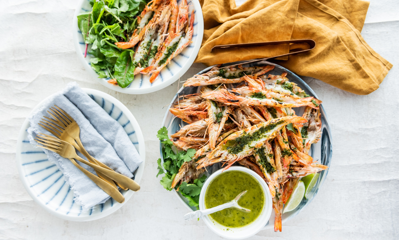 Plate of grilled shrimp garnished with herbs, accompanied by a green dipping sauce, a salad plate, a cloth napkin, and a set of gold cutlery on a table