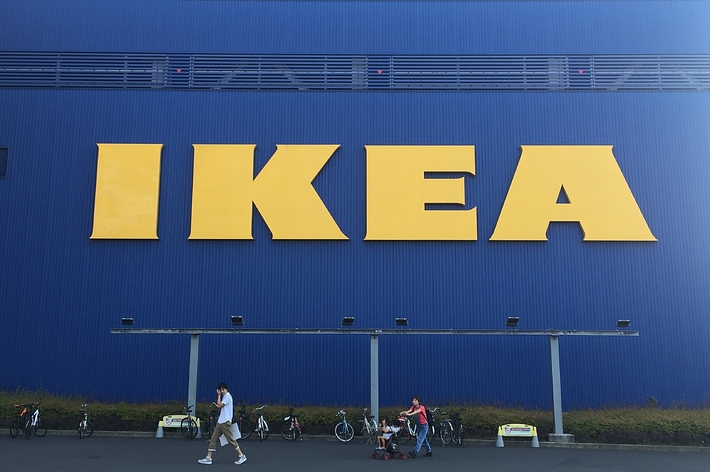 数名の人々がIKEAの店舗前を歩いている。巨大な黄色のIKEAロゴが建物の側面に見える。