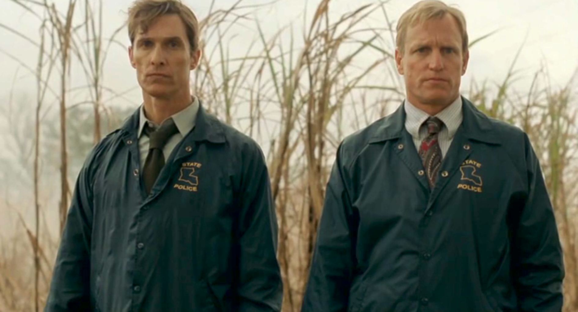 Matthew McConaughey and Woody Harrelson, dressed as police officers, stand in a field with tall grass