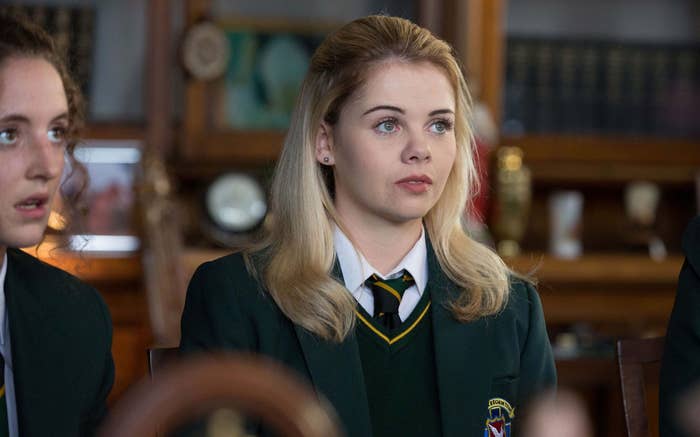 Saoirse-Monica Jackson, dressed in a school uniform with a blazer and tie, sits attentively in a classroom scene from "Derry Girls."