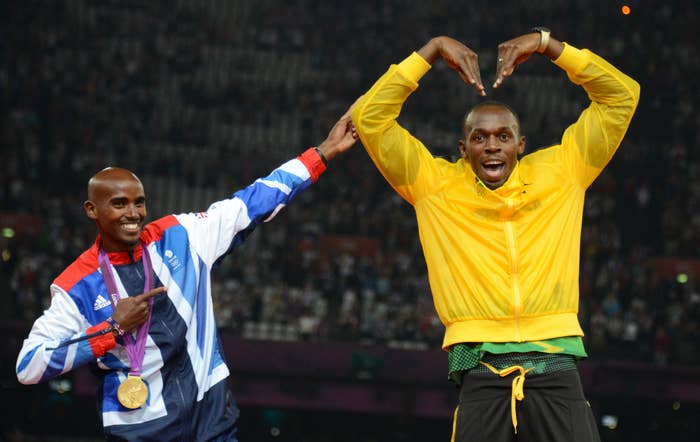 Mo Farah pointing to Usain Bolt, who is forming a heart shape with his arms above his head. Both are smiling and wearing athletic gear at a track and field event