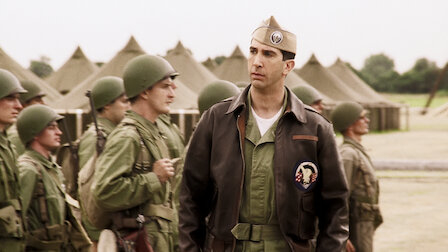 Several soldiers in military uniforms and helmets stand in formation. A man, David Schwimmer, wearing a leather jacket and a peaked cap, stands in front