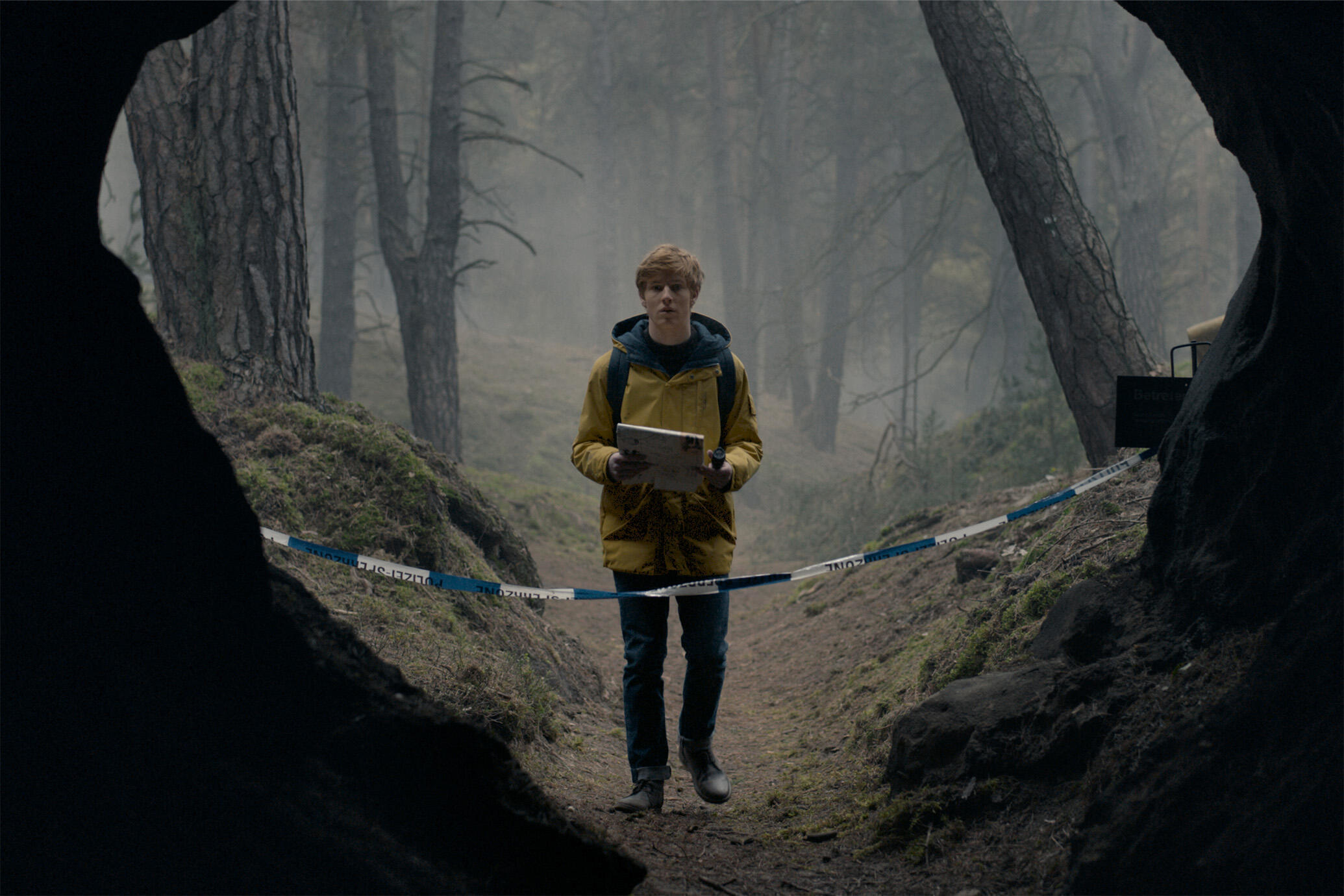 Jonas Kahnwald from "Dark" stands in a foggy forest, holding a journal. He is wearing a yellow coat with blue jeans, near police tape blocking a cave entrance