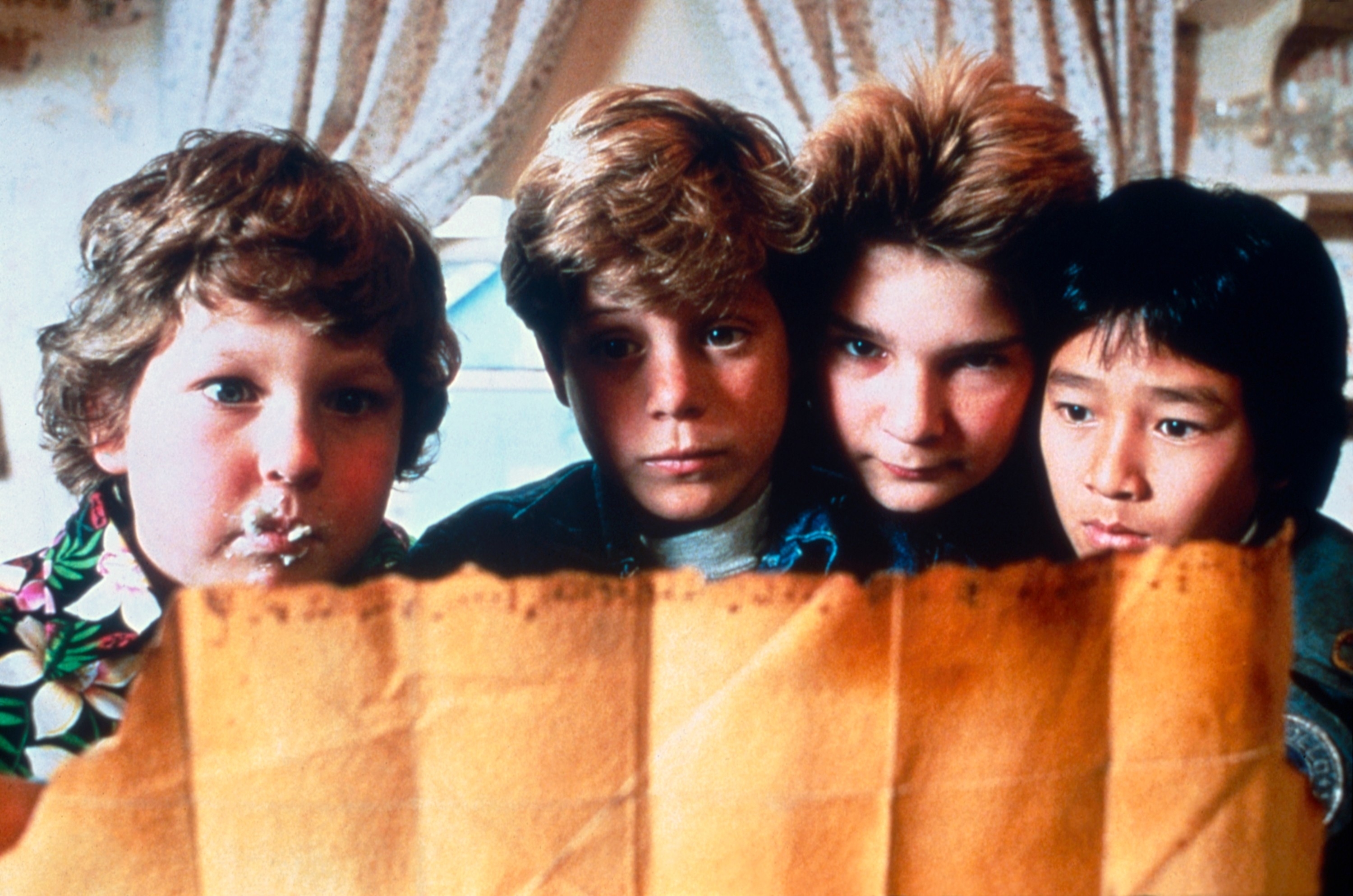 Four young boys, Jeff Cohen, Sean Astin, Corey Feldman, and Jonathan Ke Quan, intensely examine an old, brown, tattered map