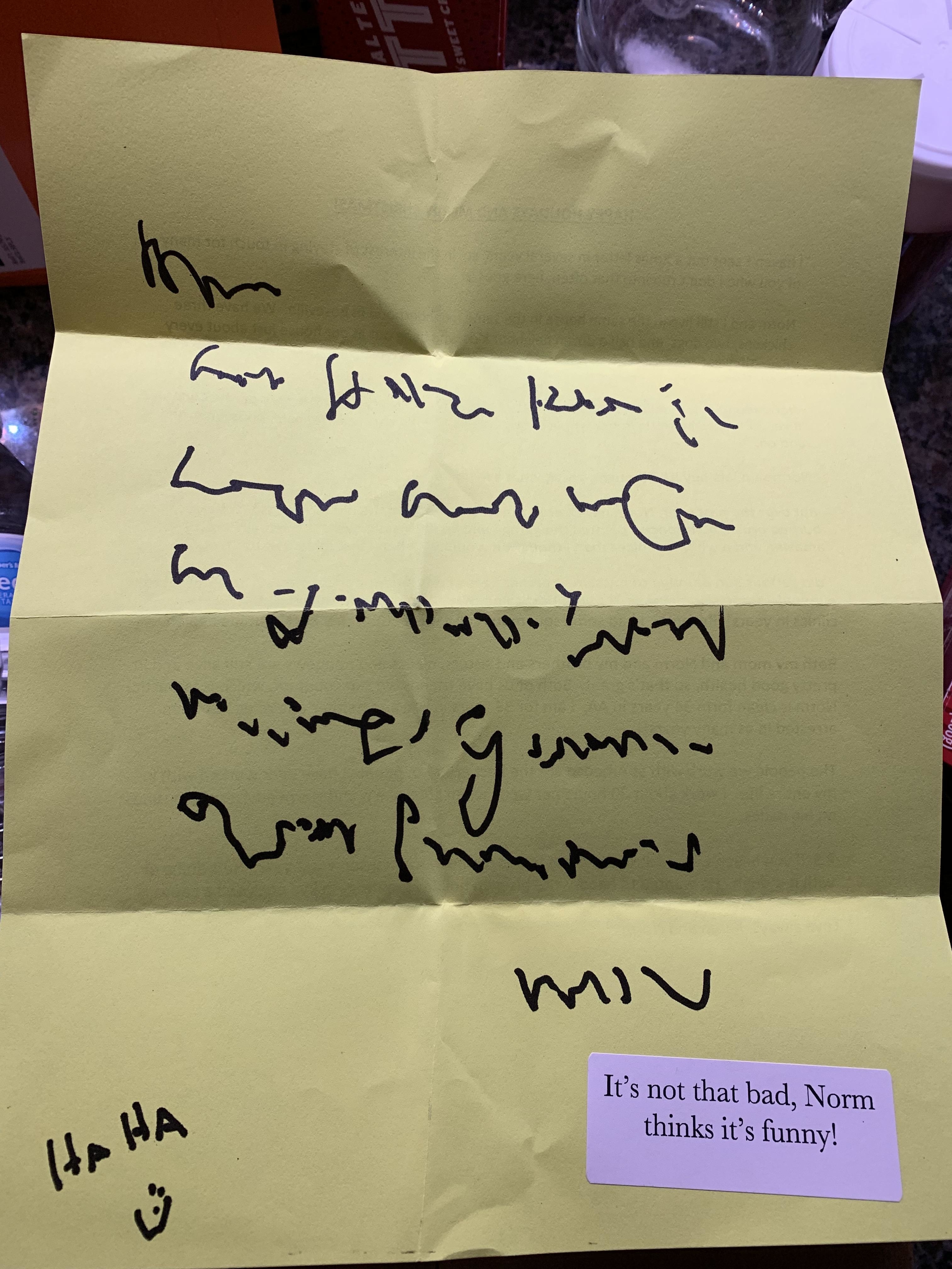 A handwritten yellow note with unreadable text. A printed label at the bottom says, "It’s not that bad, Norm thinks it’s funny!" The corner has "Ha Ha" with a smiley face