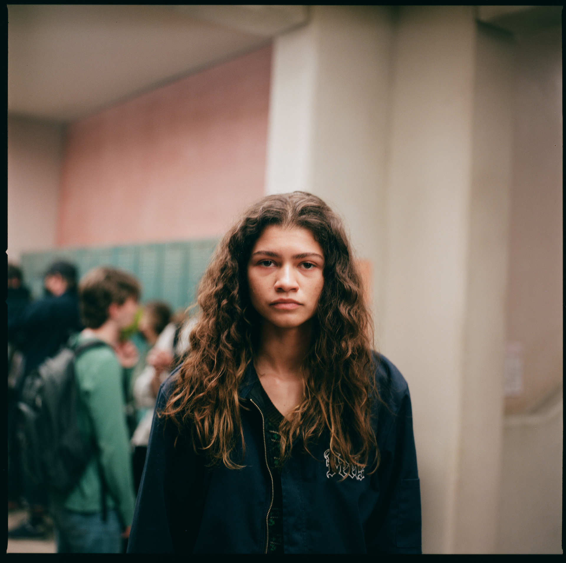 Zendaya standing in a school hallway, wearing a casual dark sweatshirt, with students blurred in the background