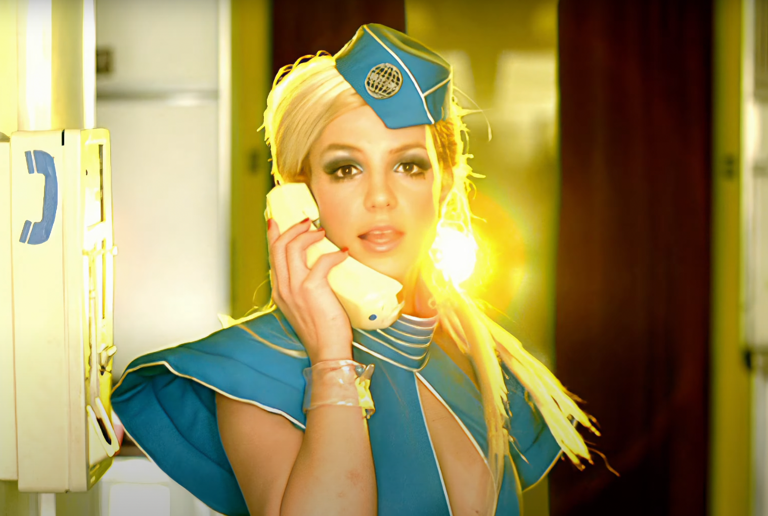 Britney Spears dressed as a flight attendant, holding a phone handset to her ear, with yellow light in the background