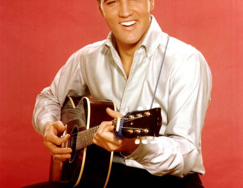 Elvis Presley plays a guitar while smiling, wearing a button-up shirt and dark pants in front of a plain backdrop