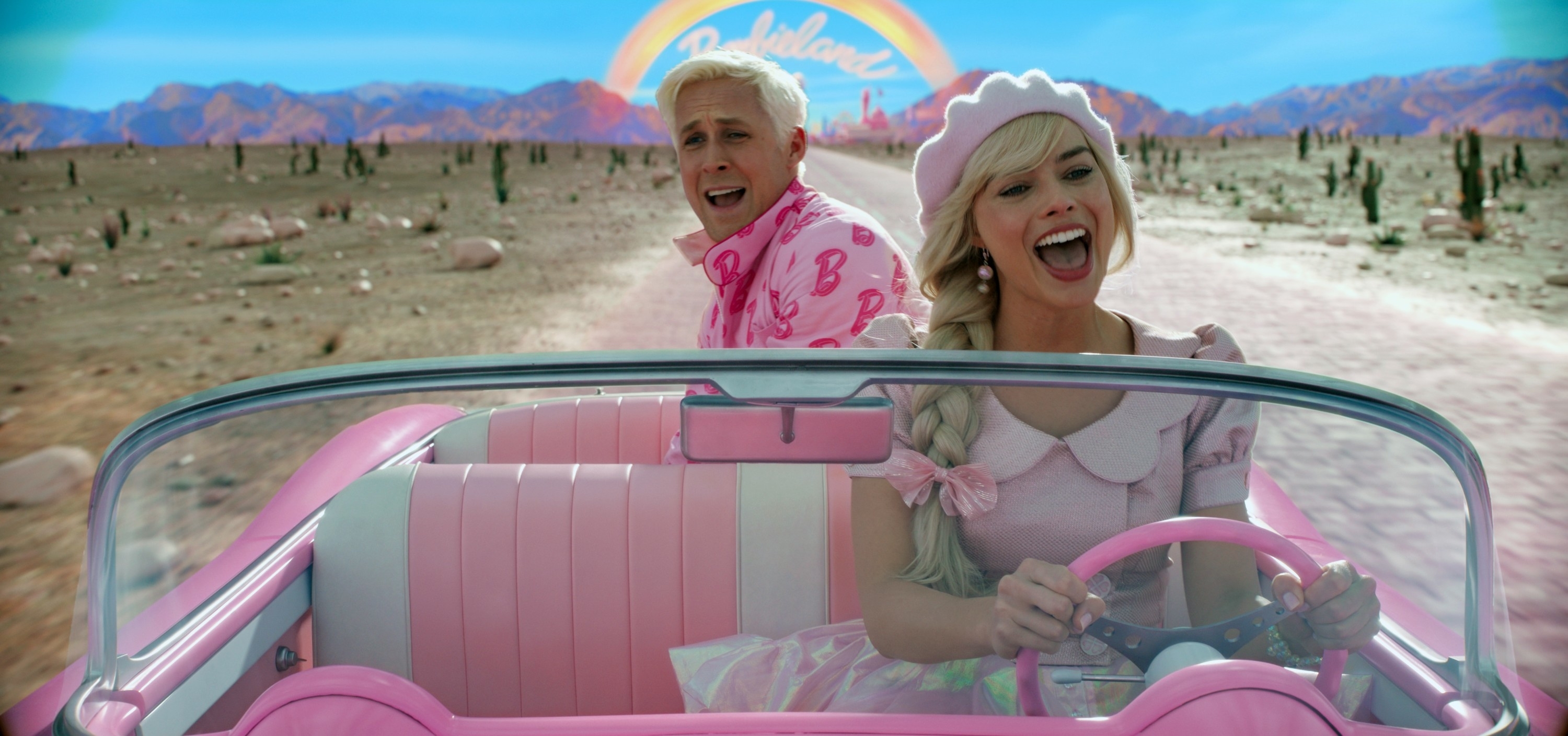 Ryan Gosling and Margot Robbie dressed in pink, driving a pink convertible in a desert with cacti and a rainbow &quot;Barbie&quot; sign in the background
