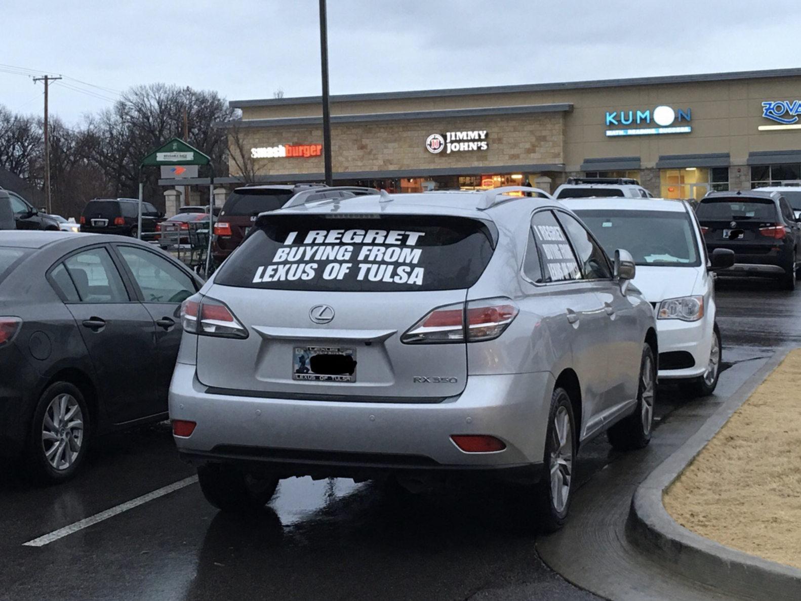 A silver Lexus SUV with a large sign on its rear window that reads, "I REGRET BUYING FROM LEXUS OF TULSA," parked in a grocery store parking lot