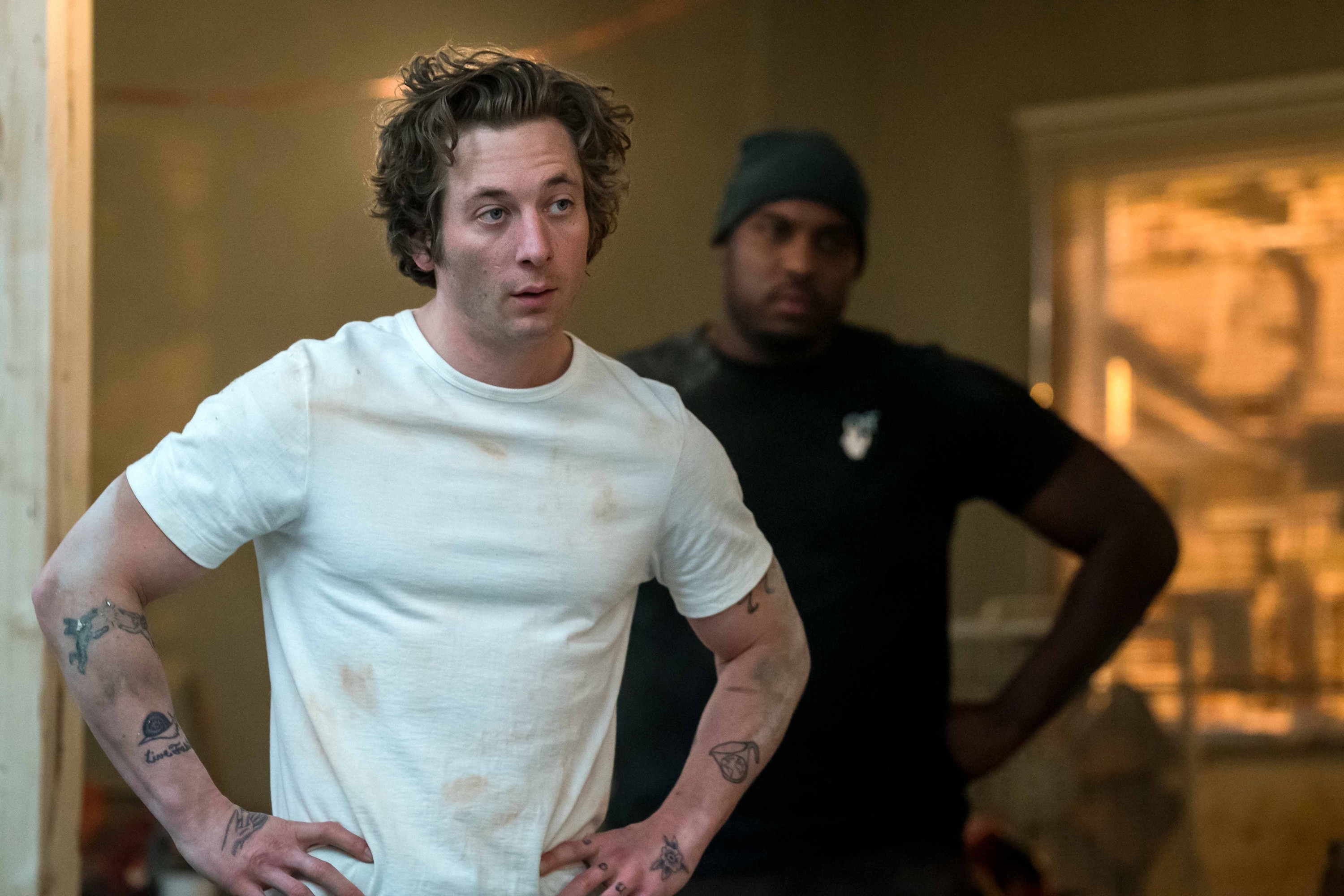 Jeremy Allen White in a dirty white t-shirt stands with hands on hips. Another person in the background watches