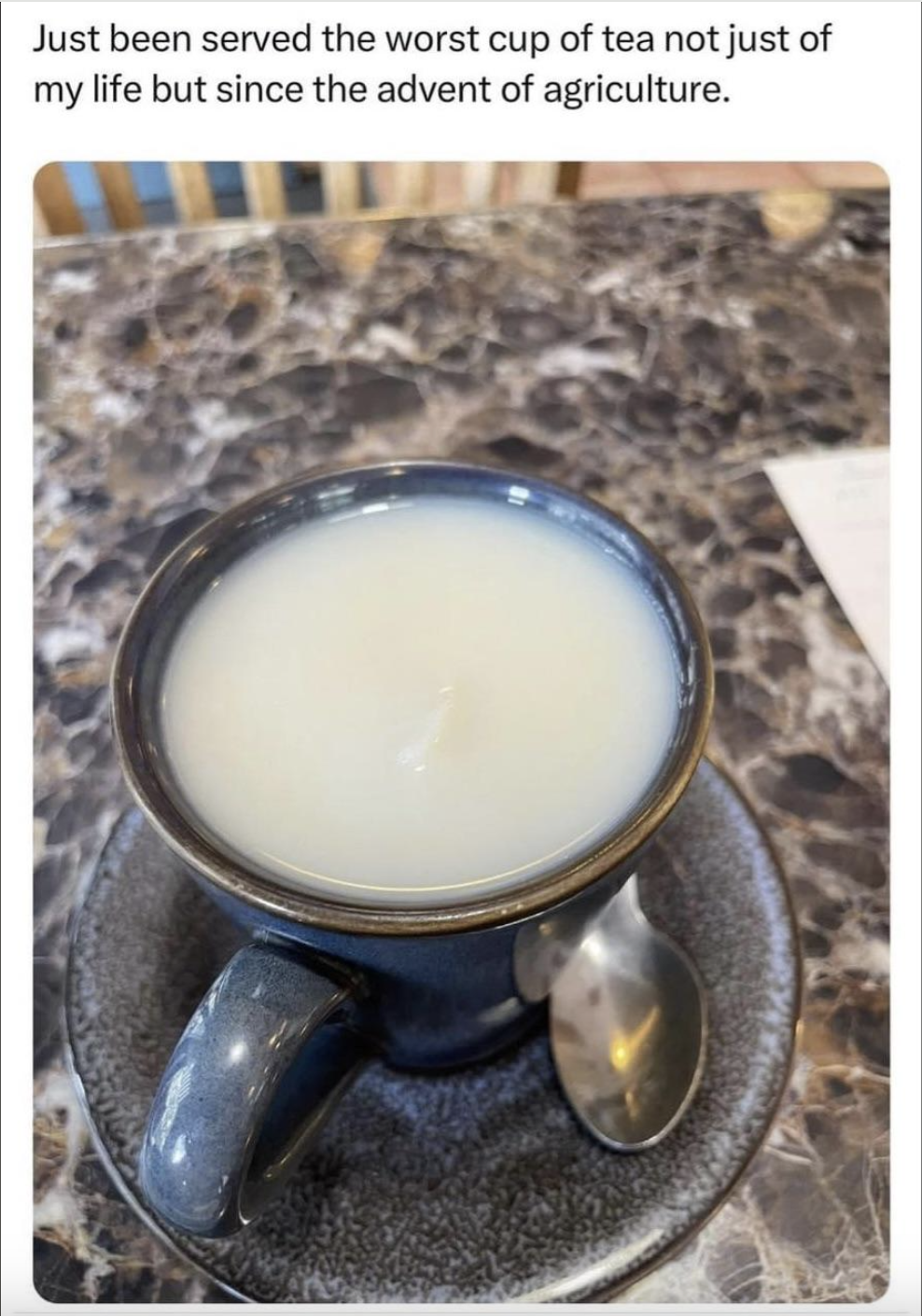 A poorly mixed cup of tea with an excessive amount of milk, on a marble table with a spoon on the saucer. Text above reads: "Just been served the worst cup of tea not just of my life but since the advent of agriculture."