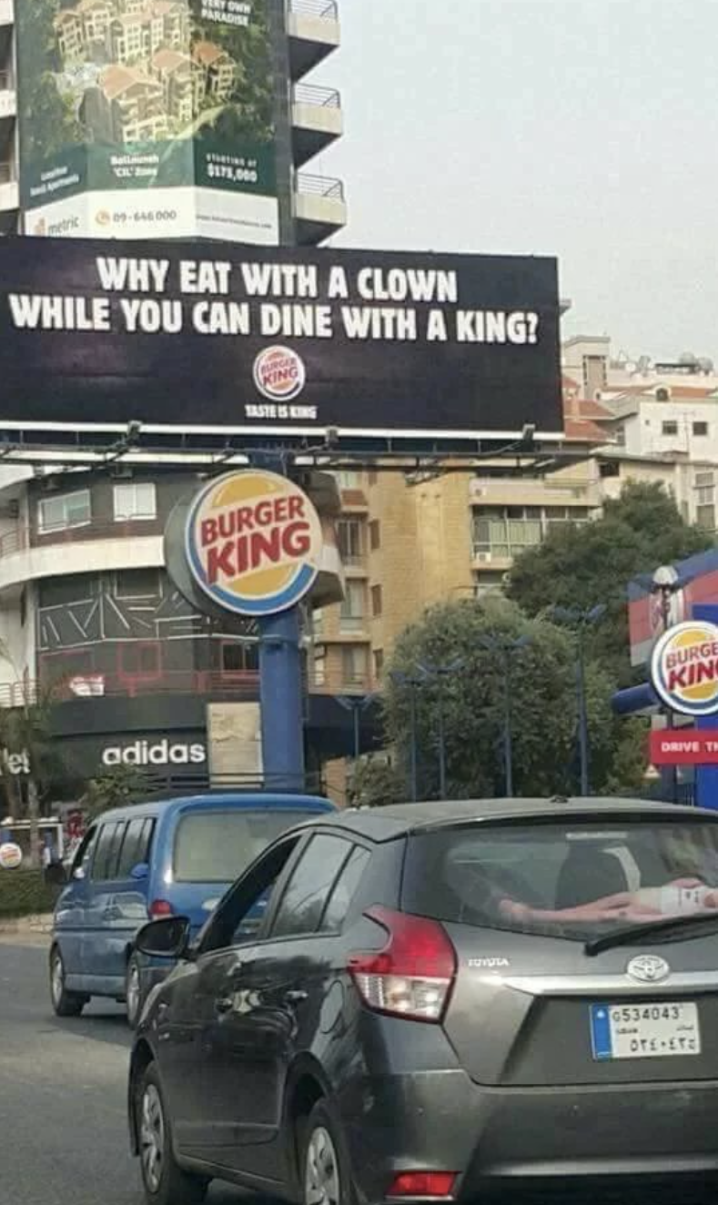 Burger King billboard in a city reads, "Why eat with a clown when you can dine with a king?" Cars are driving past the sign