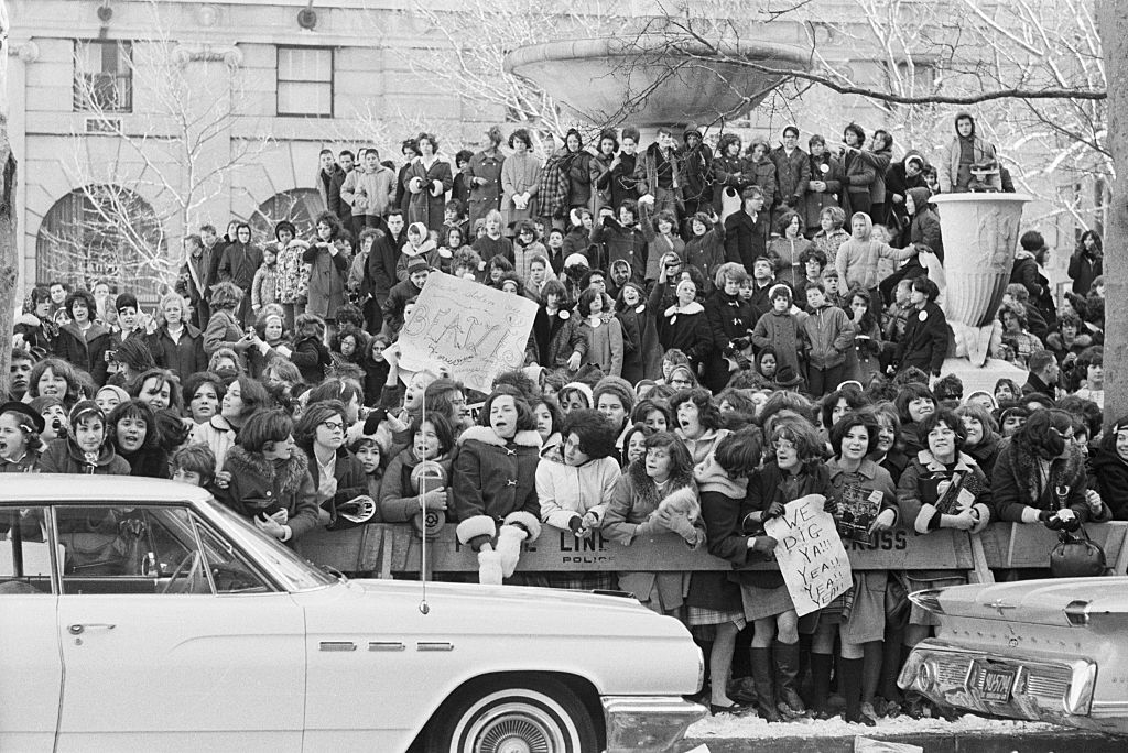 A large crowd gathers outside a building, with some people holding up signs. There are two classic cars in the foreground