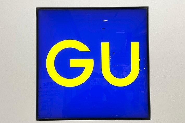 A square sign with the letters “GU” in yellow on a blue background. The sign has a plain background