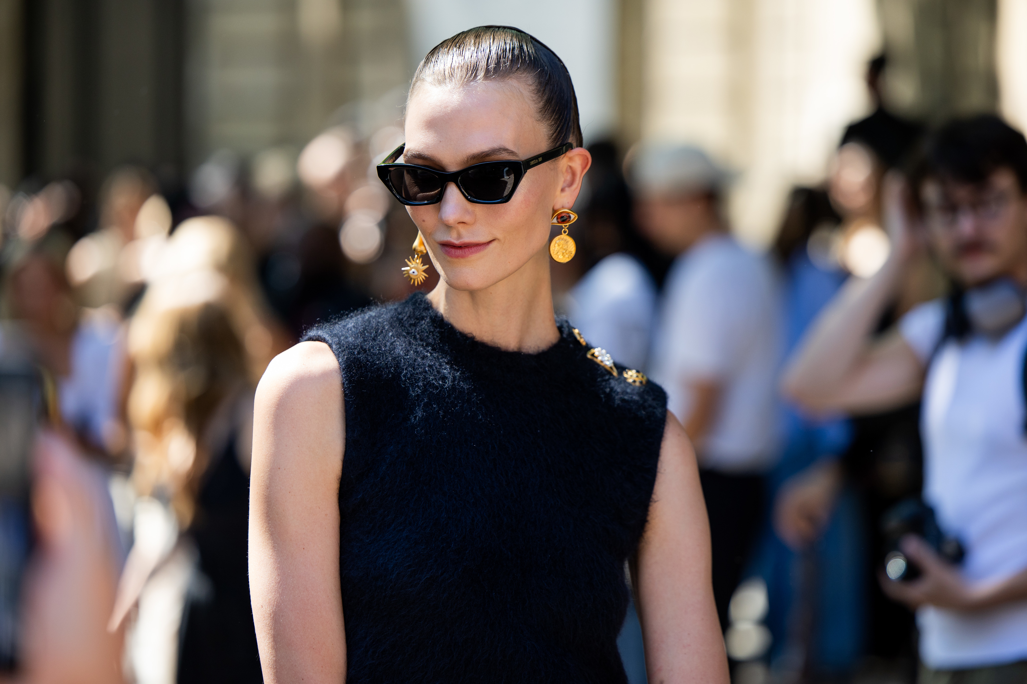 Karlie Kloss wearing a sleeveless black top, stylish sunglasses, and gold star-shaped earrings, posing outdoors amidst a crowd