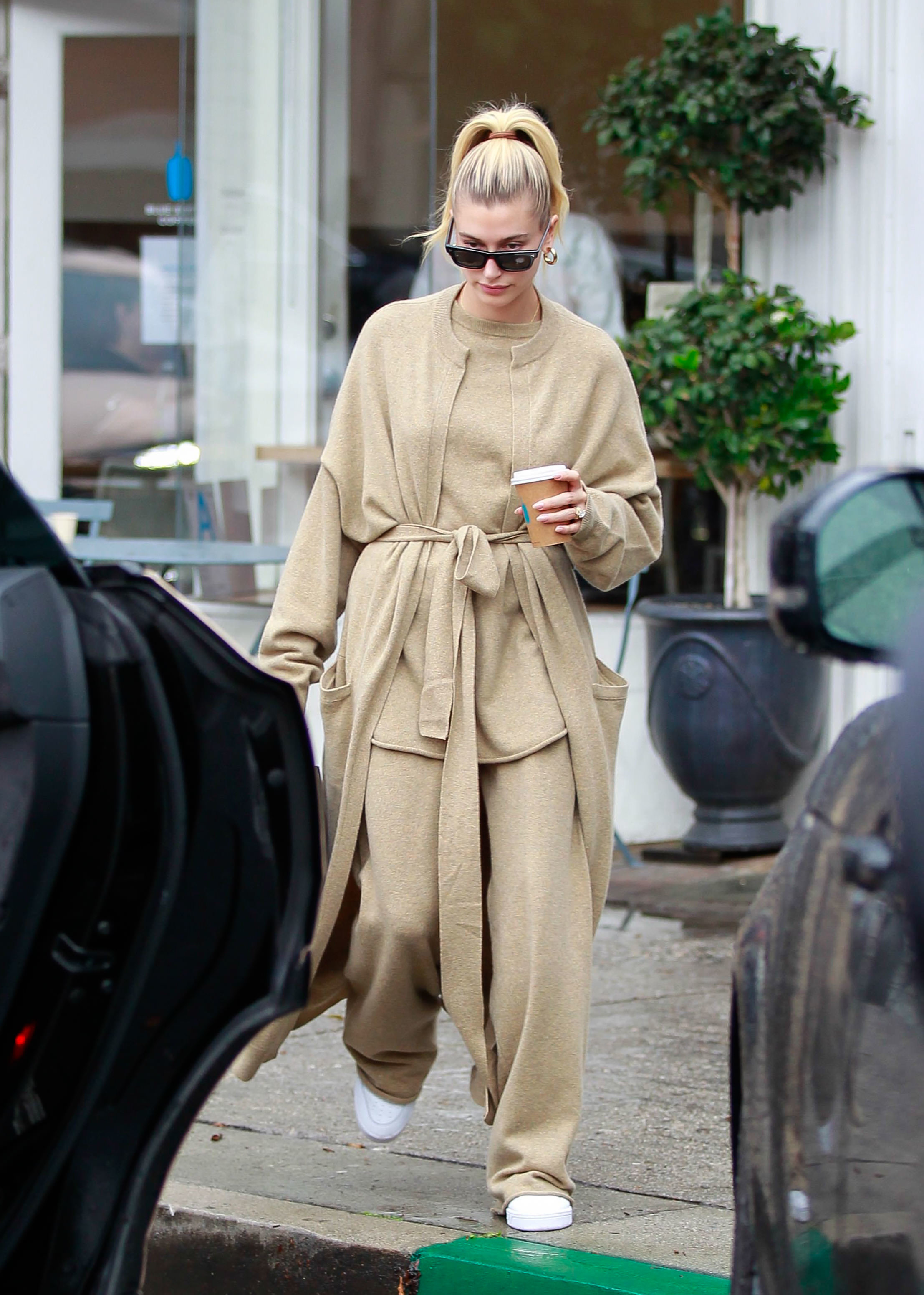 Hailey Bieber sai com uma roupa bege casual e grande, segurando uma xícara de café e usando óculos escuros, enquanto caminha na calçada