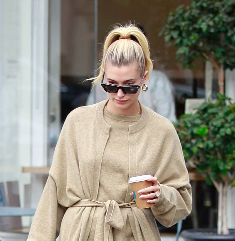 Hailey Bieber sai com uma roupa bege casual e grande, segurando uma xícara de café e usando óculos escuros, enquanto caminha na calçada