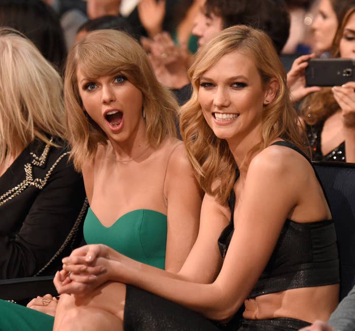 Taylor Swift in a strapless dress and Karlie Kloss in a sleeveless dress sitting together, smiling and looking excited at an event