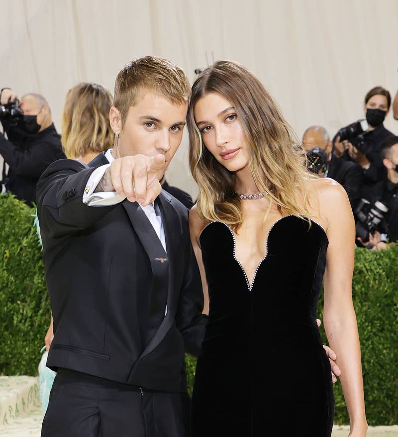 Justin Bieber de terno e tênis aponta para a câmera, ao lado de Hailey Bieber em um vestido sem alças em um evento no tapete vermelho