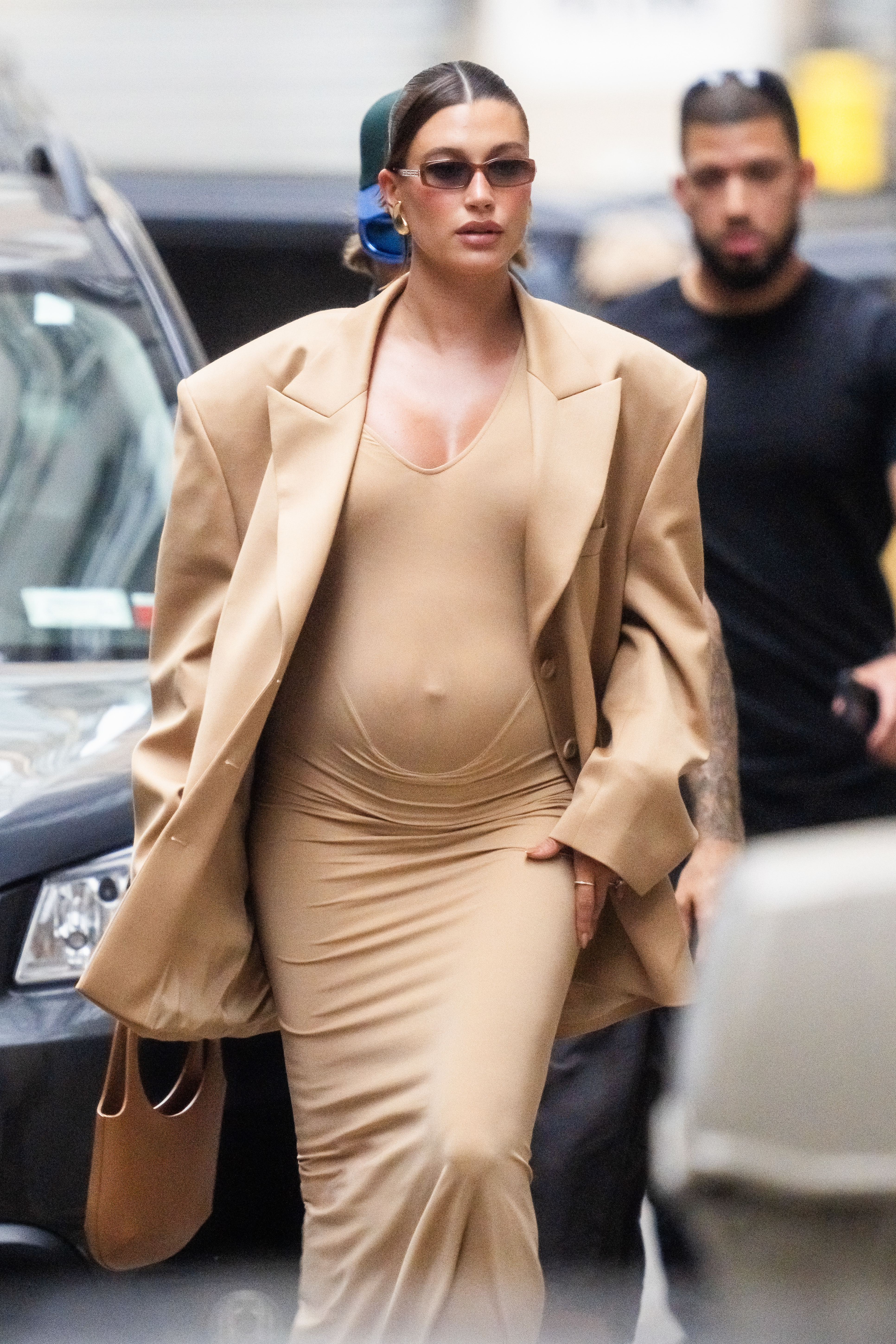 Hailey Bieber está andando na rua usando um vestido bege justo e um blazer bege grande. Ela está usando óculos escuros e seu cabelo está preso para trás. Um homem a segue