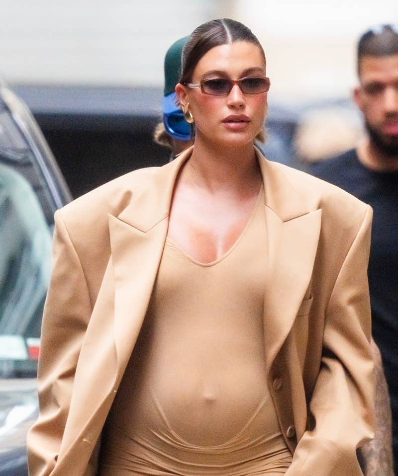 Hailey Bieber está andando na rua usando um vestido bege justo e um blazer bege grande. Ela está usando óculos escuros e seu cabelo está preso para trás. Um homem a segue