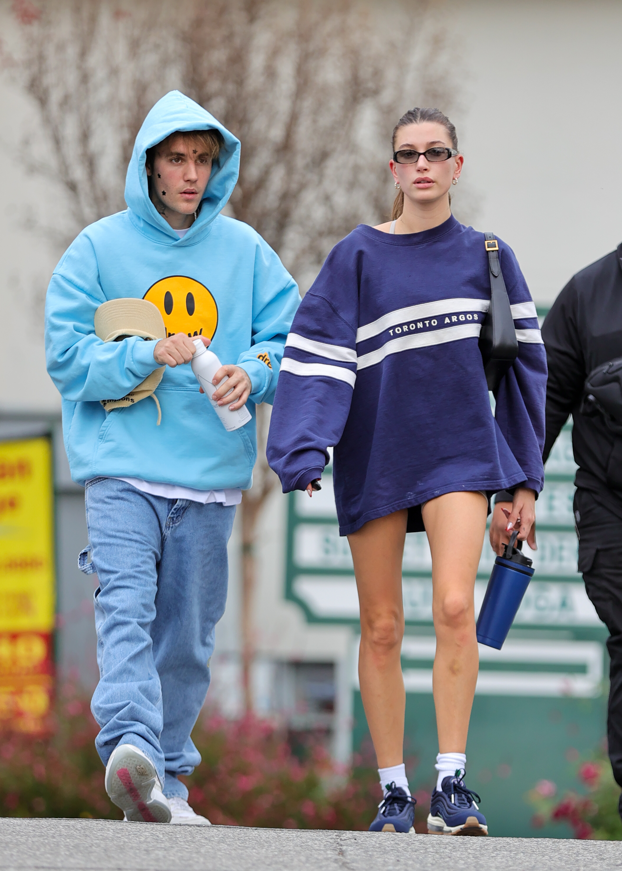 Justin Bieber e Hailey Bieber caminham juntos; Justin com um moletom com capuz e um rosto sorridente e Hailey com um suéter grande e tênis