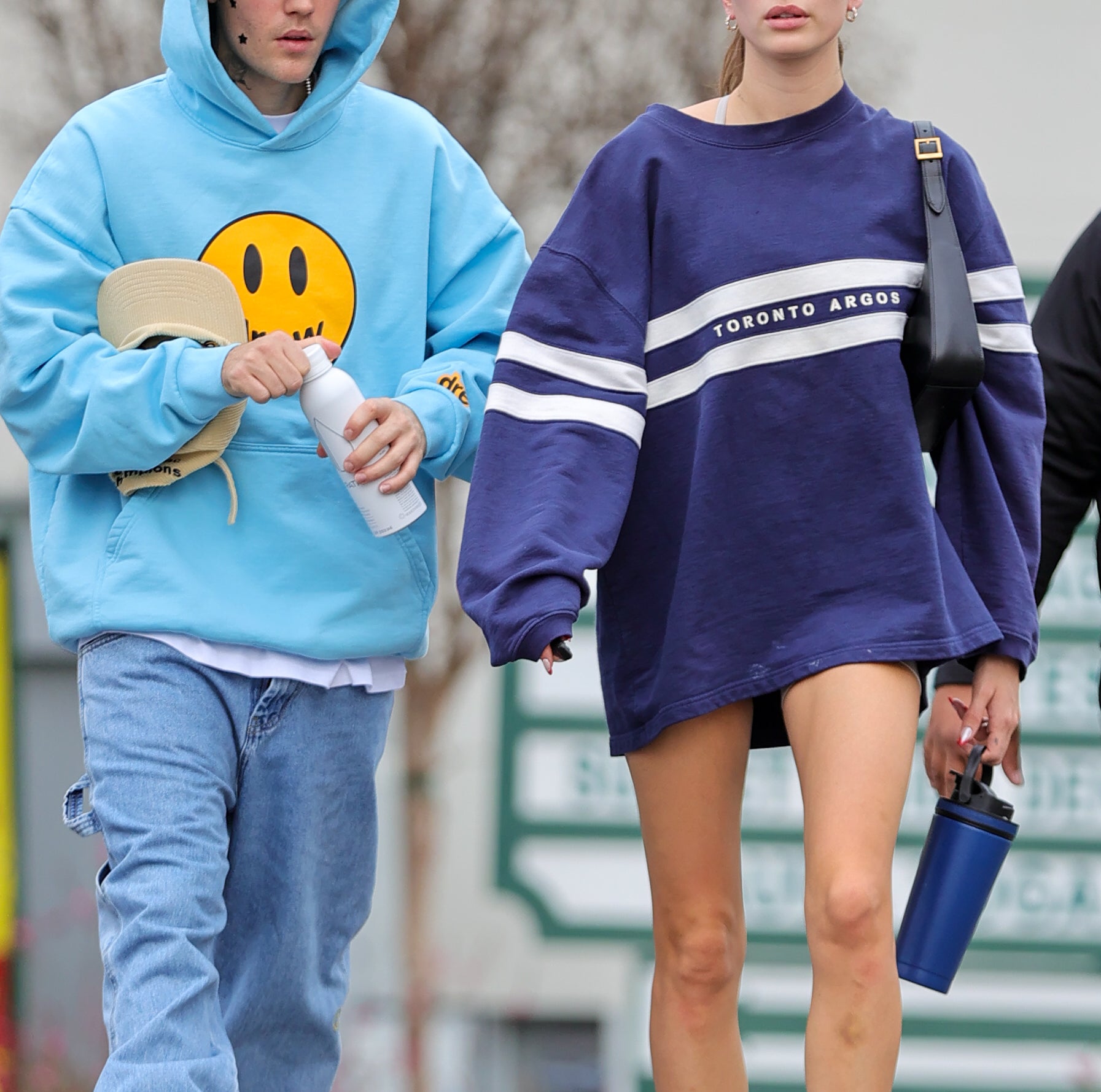 Justin Bieber and Hailey Bieber walk together; Justin in a hooded sweatshirt with a smiley face and Hailey in an oversized sweater and sneakers