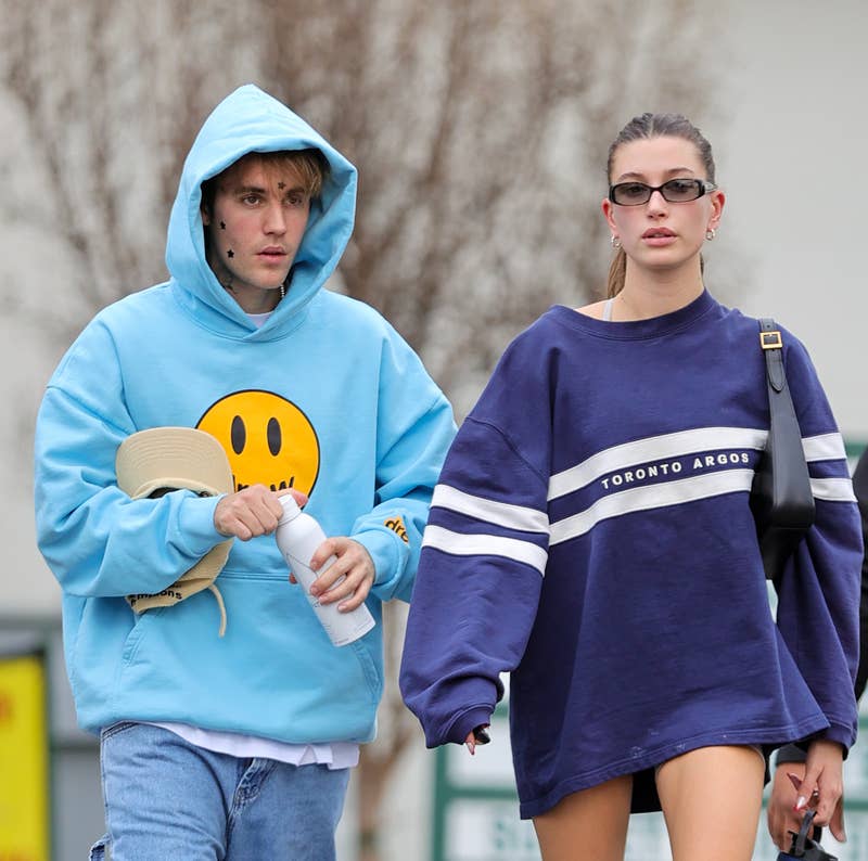 Justin Bieber e Hailey Bieber caminham juntos; Justin com um moletom com capuz e um rosto sorridente e Hailey com um suéter grande e tênis