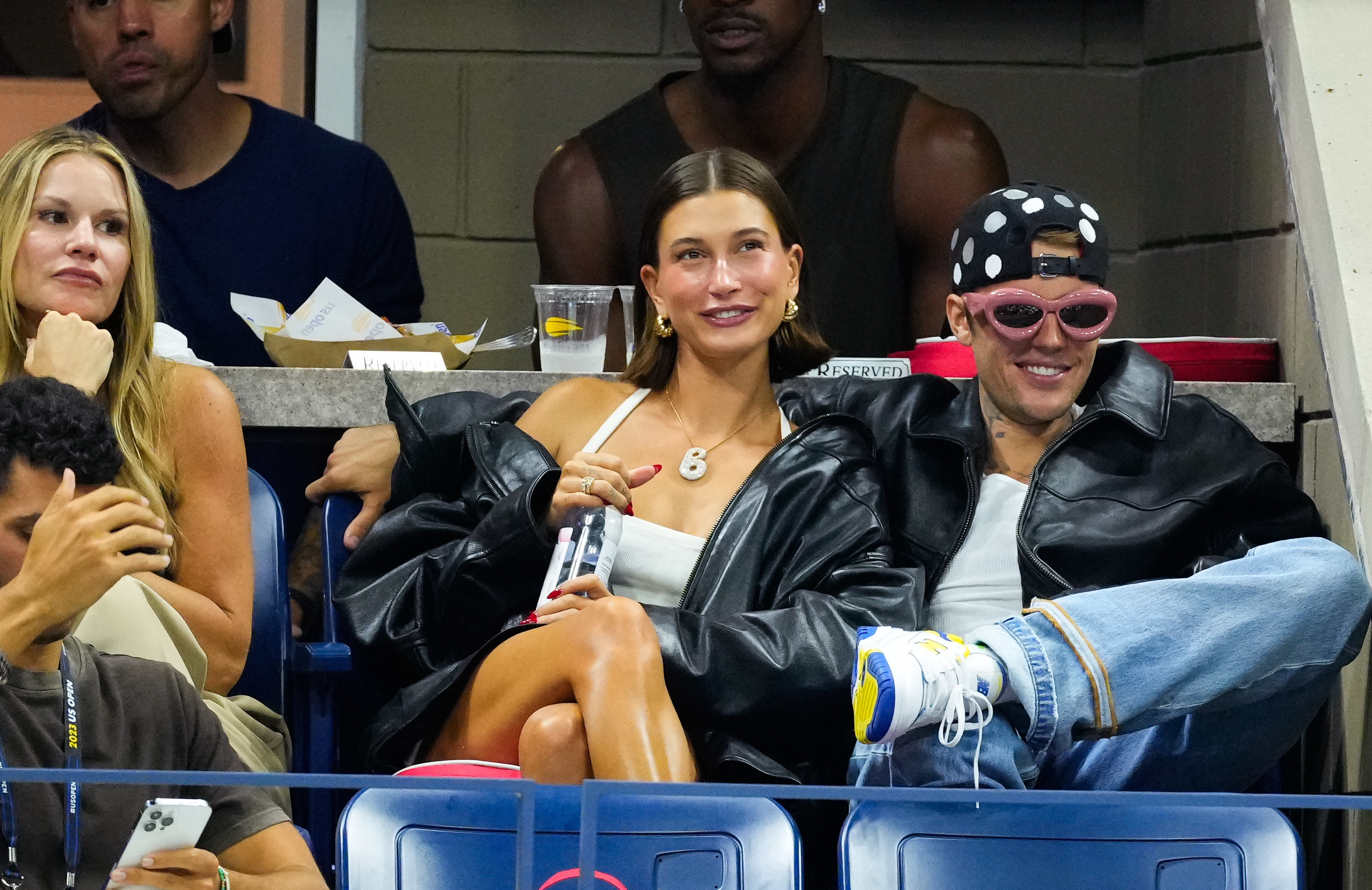 Hailey e Justin Bieber sentam-se bem próximos em um evento. Hailey está usando uma blusa branca e um colar, enquanto Justin ostenta uma jaqueta de couro, óculos de sol rosa e um chapéu de bolinhas