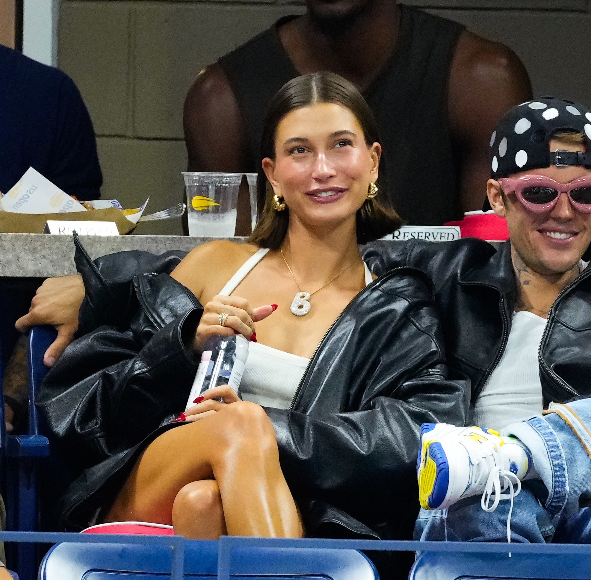 Hailey and Justin Bieber sit closely together at an event. Hailey is wearing a white top and necklace, while Justin sports a leather jacket, pink sunglasses, and a polka-dot hat