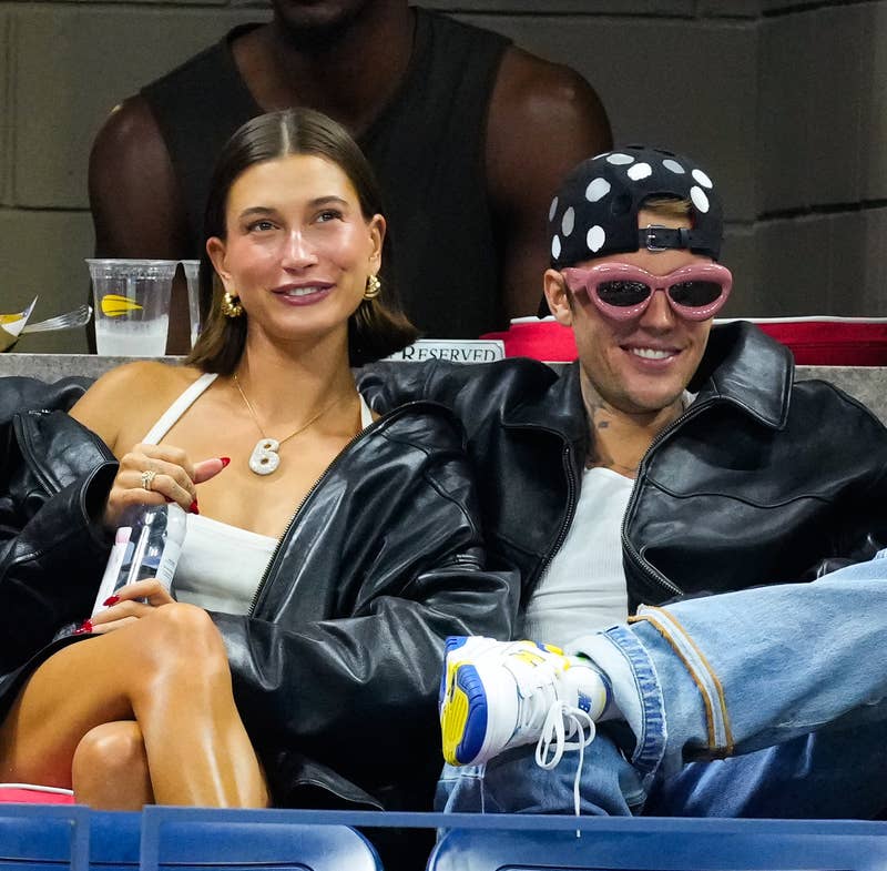 Hailey e Justin Bieber sentam-se bem próximos em um evento. Hailey está usando uma blusa branca e um colar, enquanto Justin ostenta uma jaqueta de couro, óculos de sol rosa e um chapéu de bolinhas