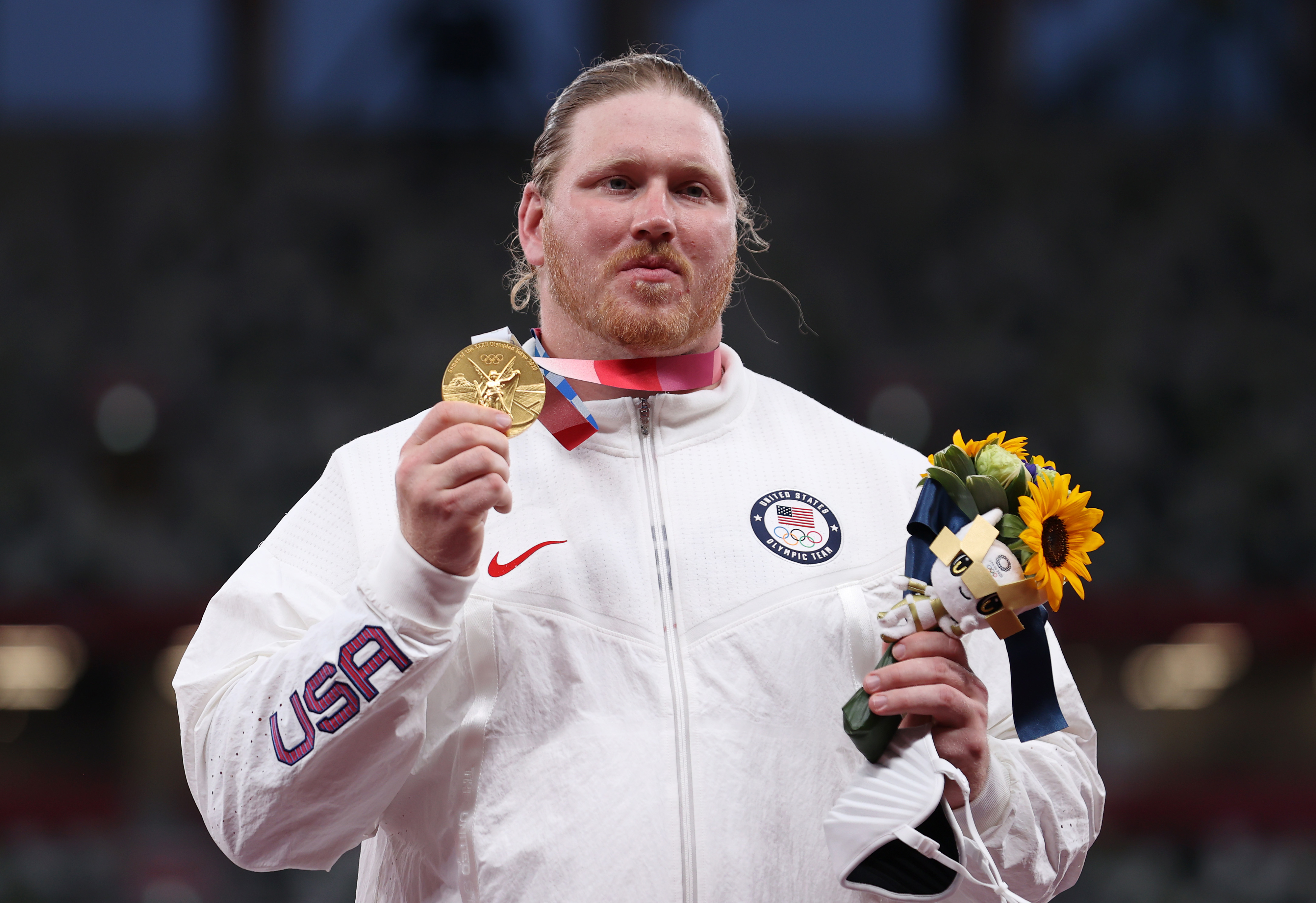 Man in a Team USA jacket holds a gold medal and a bouquet of flowers, likely on a victory podium during an official ceremony. Names unknown