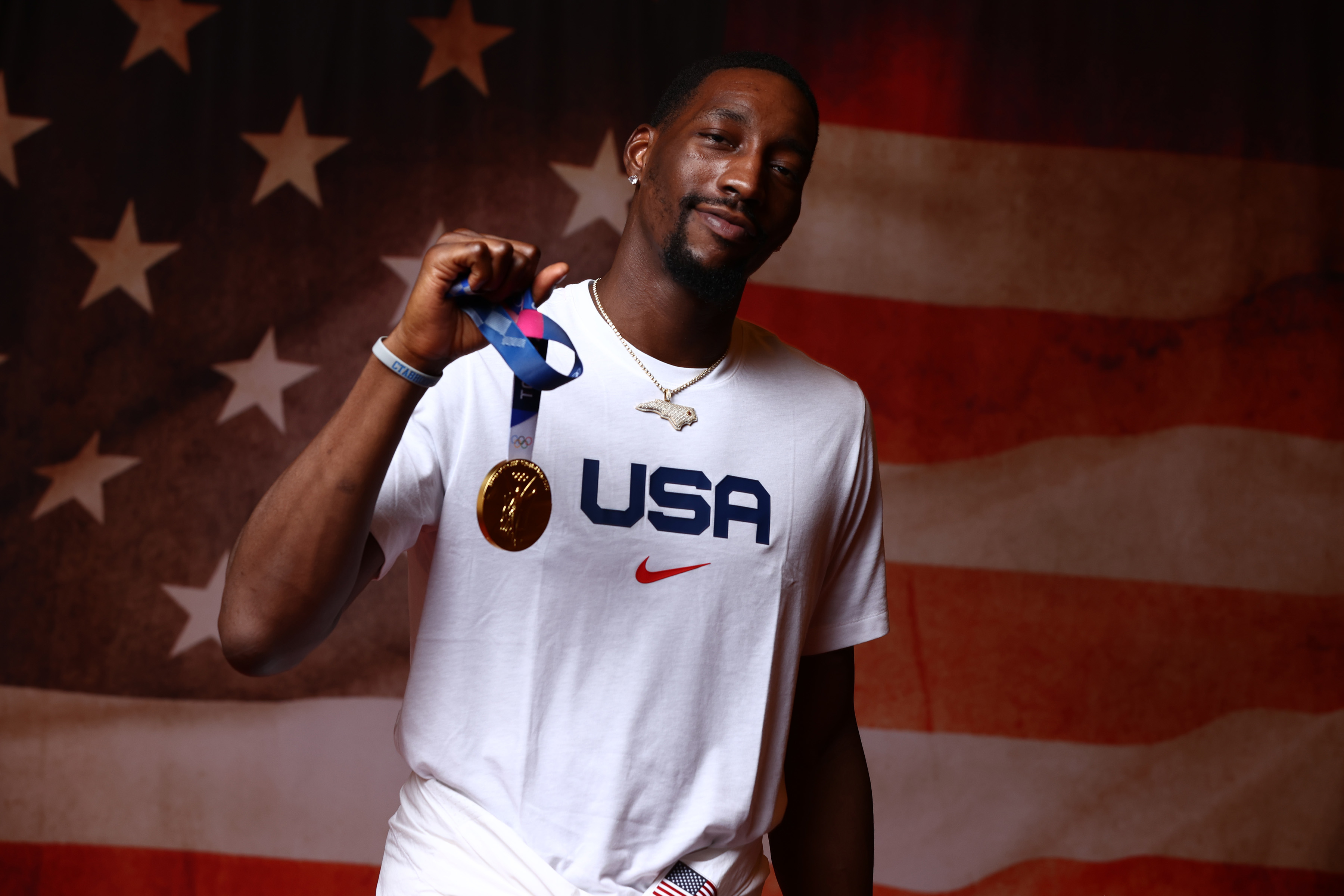 Bam Adebayo stands in front of an American flag background while holding up a gold medal, wearing a white &quot;USA&quot; T-shirt and a silver necklace