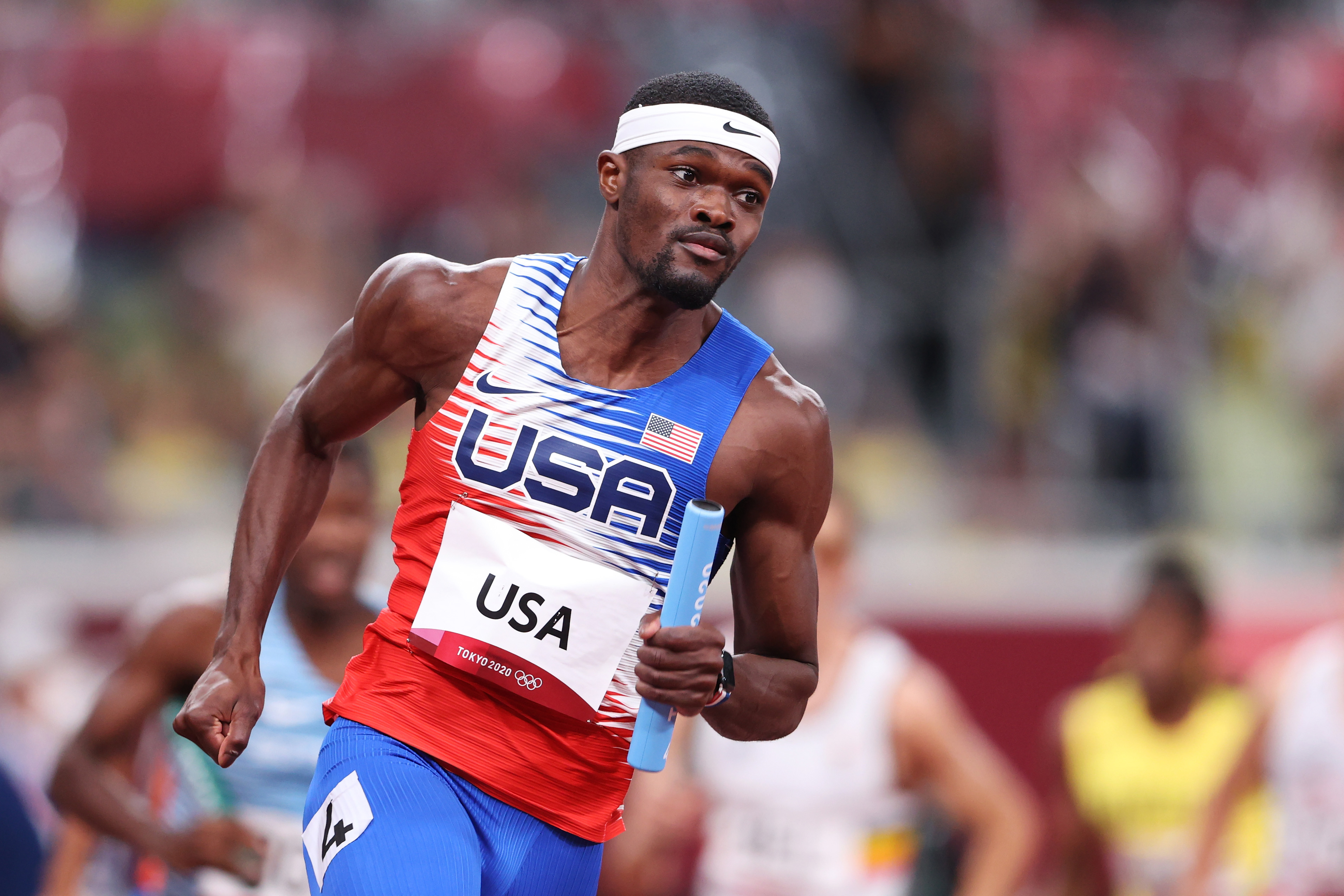 Rai Benjamin in uniform with &quot;USA&quot; text and flag, running with a relay baton in a stadium during a track event