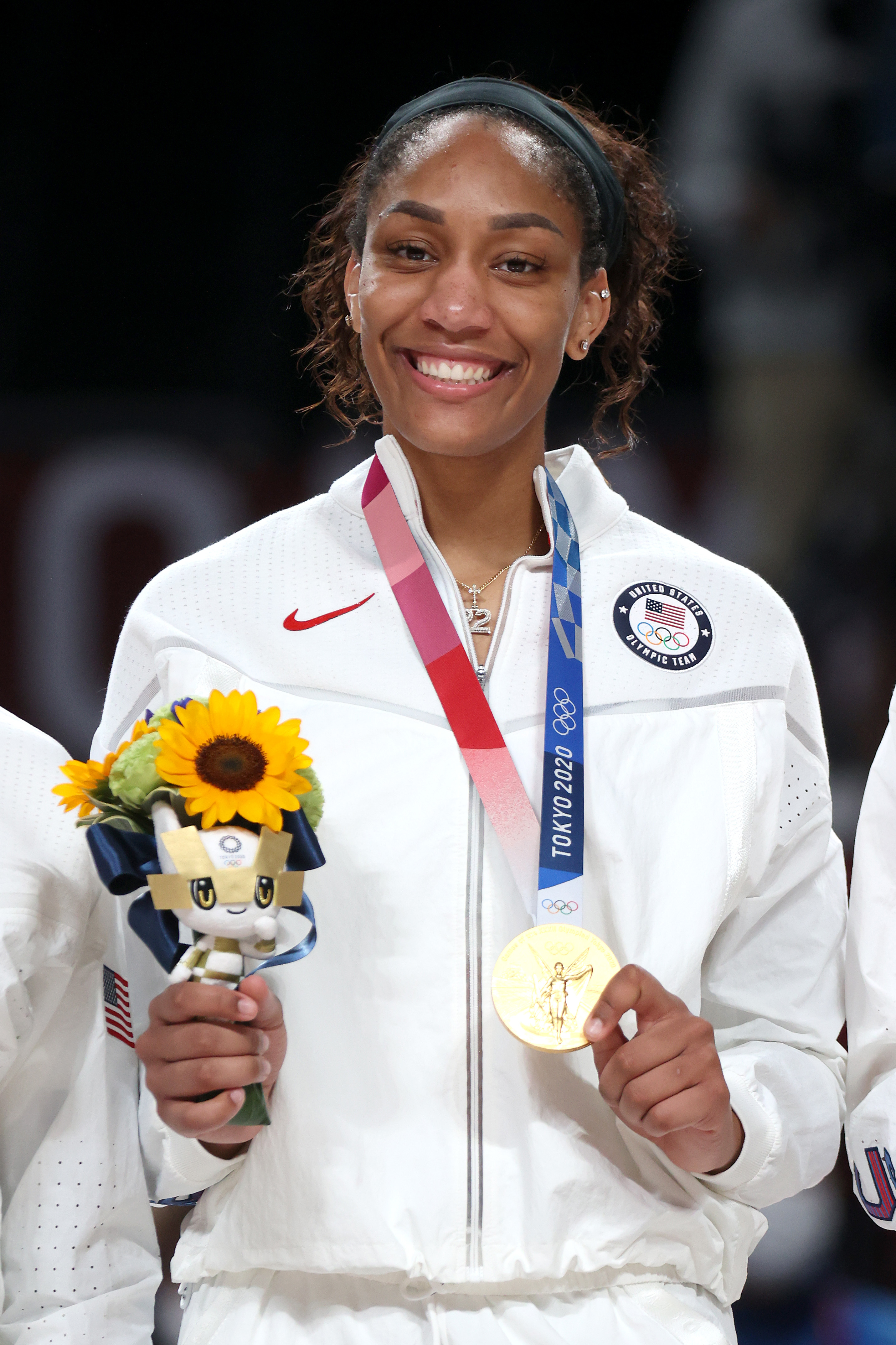 A'ja Wilson, in a USA tracksuit, smiles while holding a gold medal and flowers.