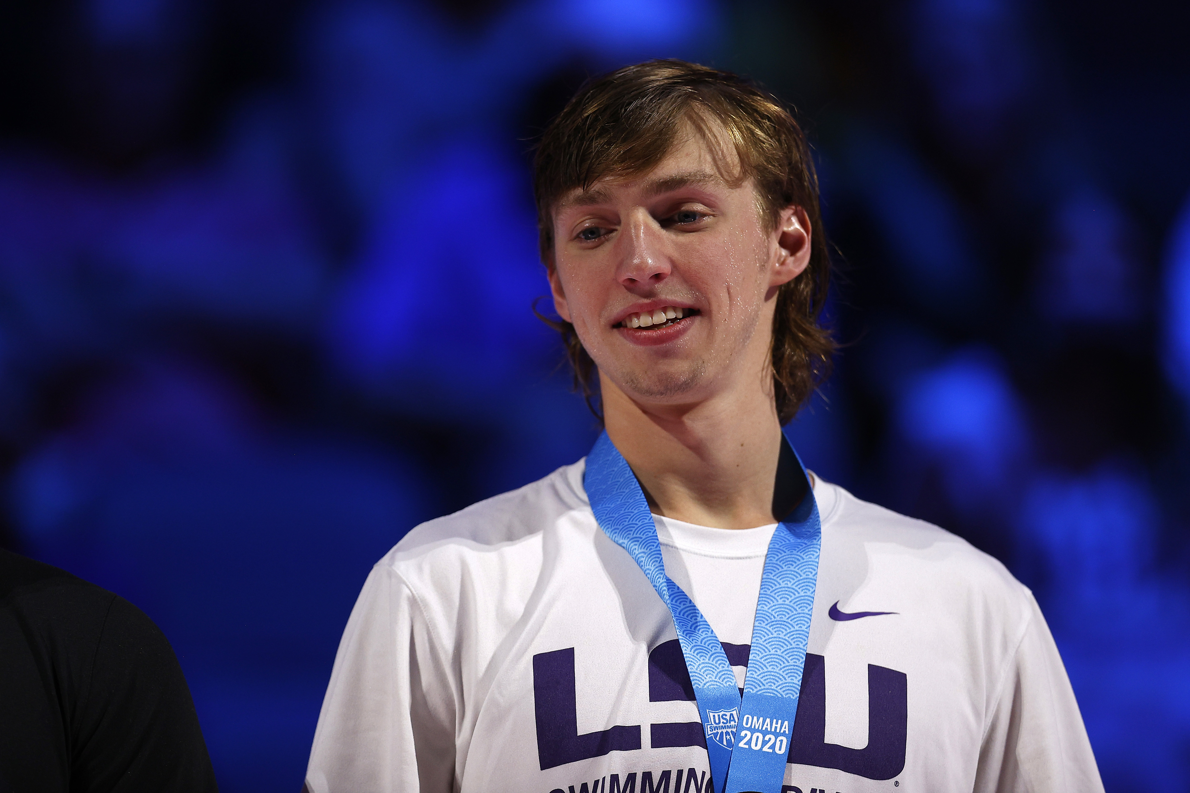Brooks Curry with a light shirt and medal around his neck stands in front of a blurred, blue-lit background