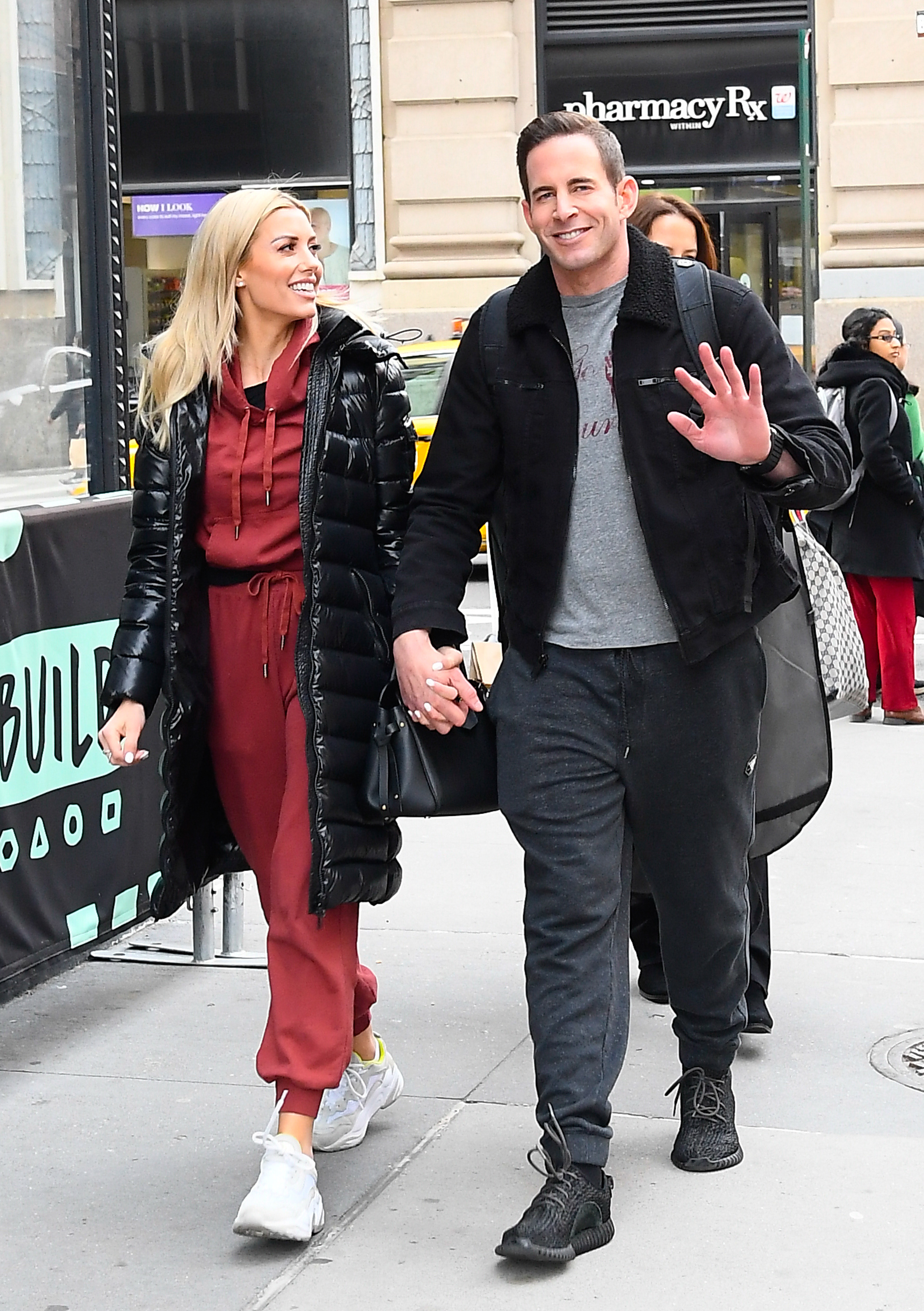 Heather Rae El Moussa in a casual outfit and Tarek El Moussa in a jacket and grey pants, walking and holding hands on a city street, smiling at the camera