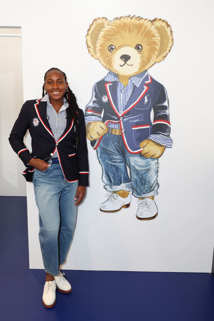Coco Gauff stands beside a large illustration of a teddy bear wearing similar clothing: a blazer, striped shirt, jeans, and white shoes