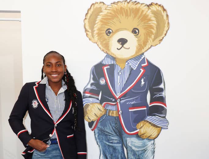 Coco Gauff stands beside a large illustration of a teddy bear wearing similar clothing: a blazer, striped shirt, jeans, and white shoes